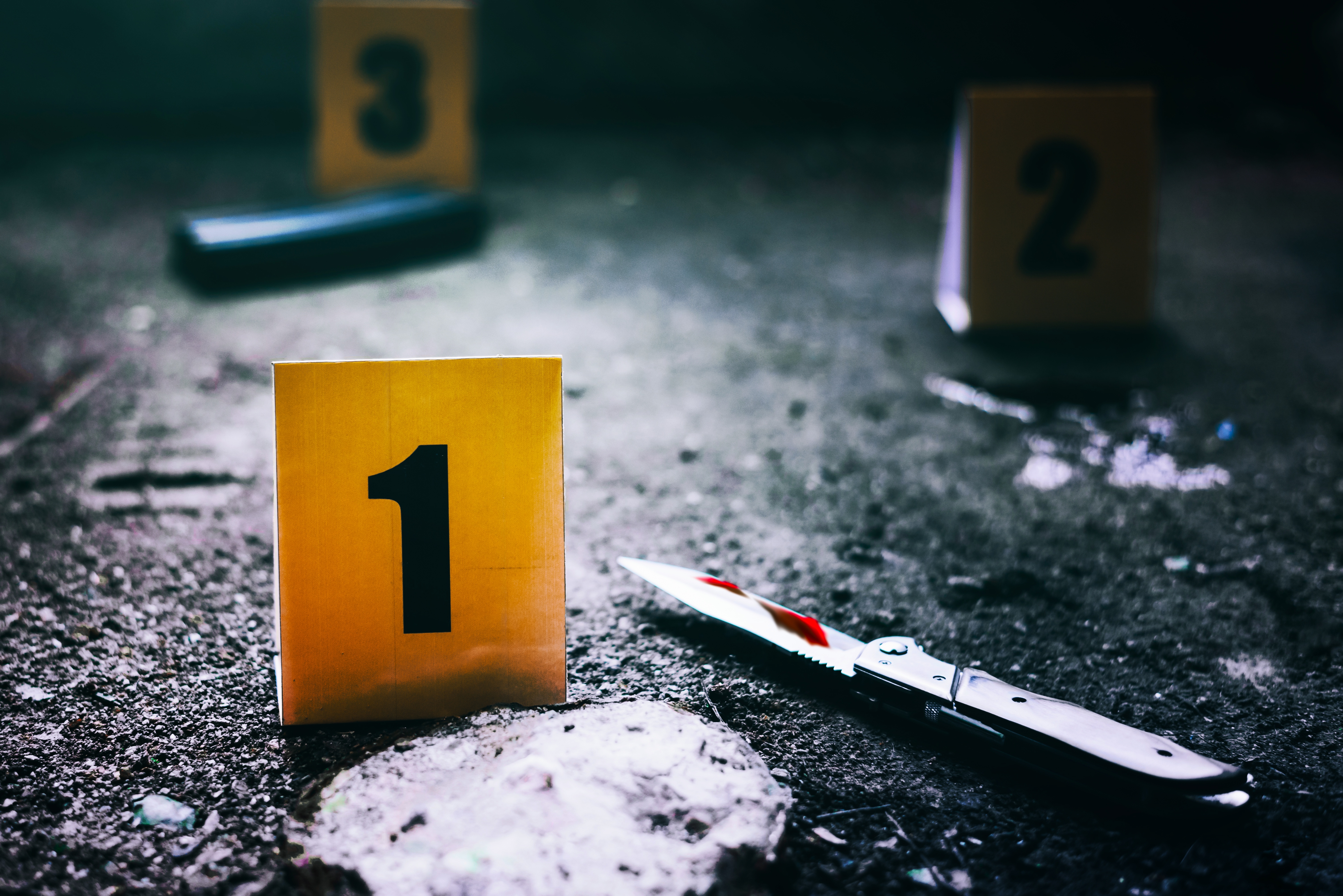 Tatort mit Spuren und Spurentafeln, im Vordergrund auf dem Boden eine kleine Spurentafel mit der schwarzen Zahl 1 und rechts daneben ein Messer mit Blutanhaftung an der Klinge, im Hintergrund unscharf zu erkennen zwei weitere gelbe Spurentafeln mit den schwarzen Zahlen 2 und 3 sowie weiteren unscharfen Spuren